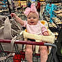 baby, infant, shopping_cart, supermarket, headband, pink_bow, bib, baby_seat, purse, packaged_food, grocery_aisle, smiling, bare_feet, legs, adult_in_background, store_signage, metal_cart, fluorescent_lighting, candid_portrait, family_shopping
