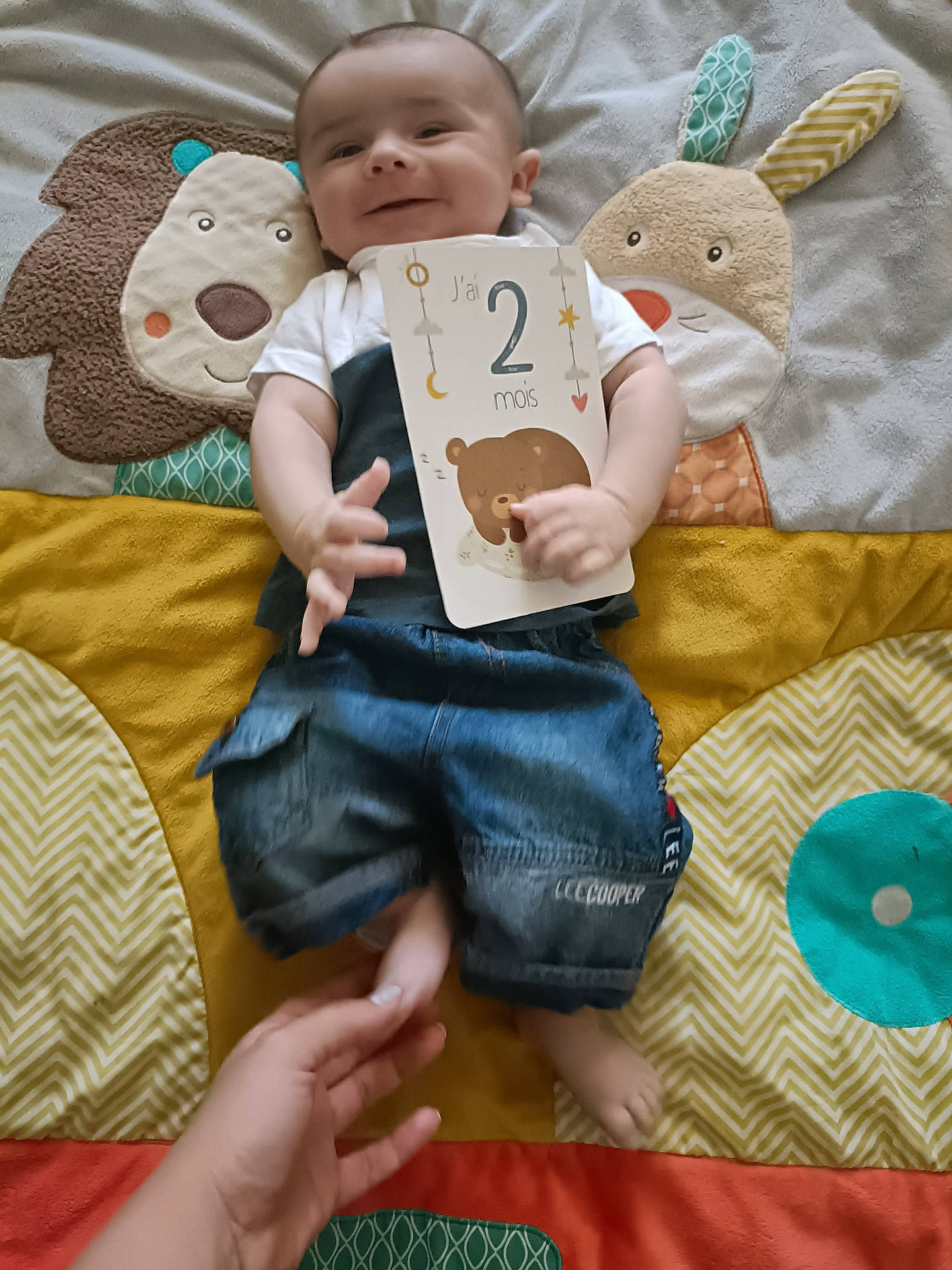 Gabin a rejoint le concours — aidez-le/la à gagner de superbes lots ! animal_design, baby, child, colorful, cute, denim_shorts, face, foot, hand, happy, indoors, infant, lying_down, milestone_card, person, play_mat, smile, soft_texture, toddler, white_shirt