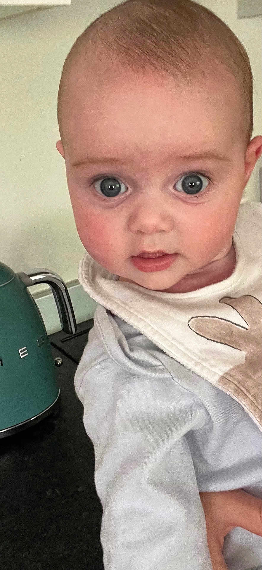 Reese is registered to the contest to win money with this photo: appliance, baby, bib, child, close_up, clothing, curious, cute, expression, eyes, face, head, indoors, infant, kettle, kitchen, person, rosy_cheeks, white, young