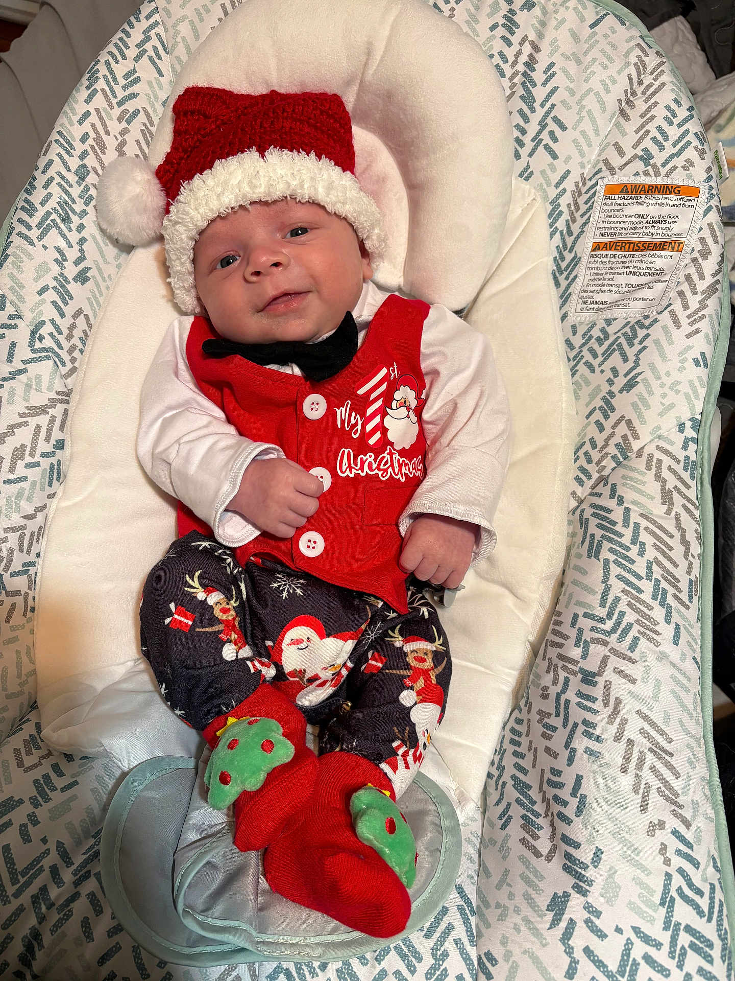 Sebastian is registered to the contest to win money with this photo: baby, infant, christmas, holiday_clothing, santa_hat, red_vest, black_bowtie, patterned_pants, christmas_socks, baby_seat, fabric_pattern, smiling, face, hands, feet, indoor, cute, festive, portrait, cozy