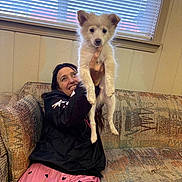 Gizmo is registered to the contest to win money with this photo: person, dog, puppy, indoor, couch, window, blinds, smiling, black_jacket, pink_pants, heart_pattern, wood_paneling, happy, pet, holding, sitting, fur, cute, cozy, home