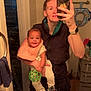 baby, person, mirror, bathroom, selfie, smile, toy, bib, towel, phone, watch, tiles, door, clothing, drawer, child, holding, green_ball, reflection, casual