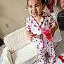 child, toddler, pajamas, hearts, smile, cup, candies, plush_chair, indoor, room, standing, rose_petals, bare_feet, clothing, portrait, happy, furniture, earrings, wall_outlet, bedspread