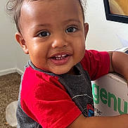 Josiah is registered to the contest to win money with this photo: child, toddler, smiling, happy, portrait, close_up, curly_hair, brown_eyes, red_shirt, gray_clothing, teeth, indoor, home, carpet, chair, sign, printed_text, arms, face, candid