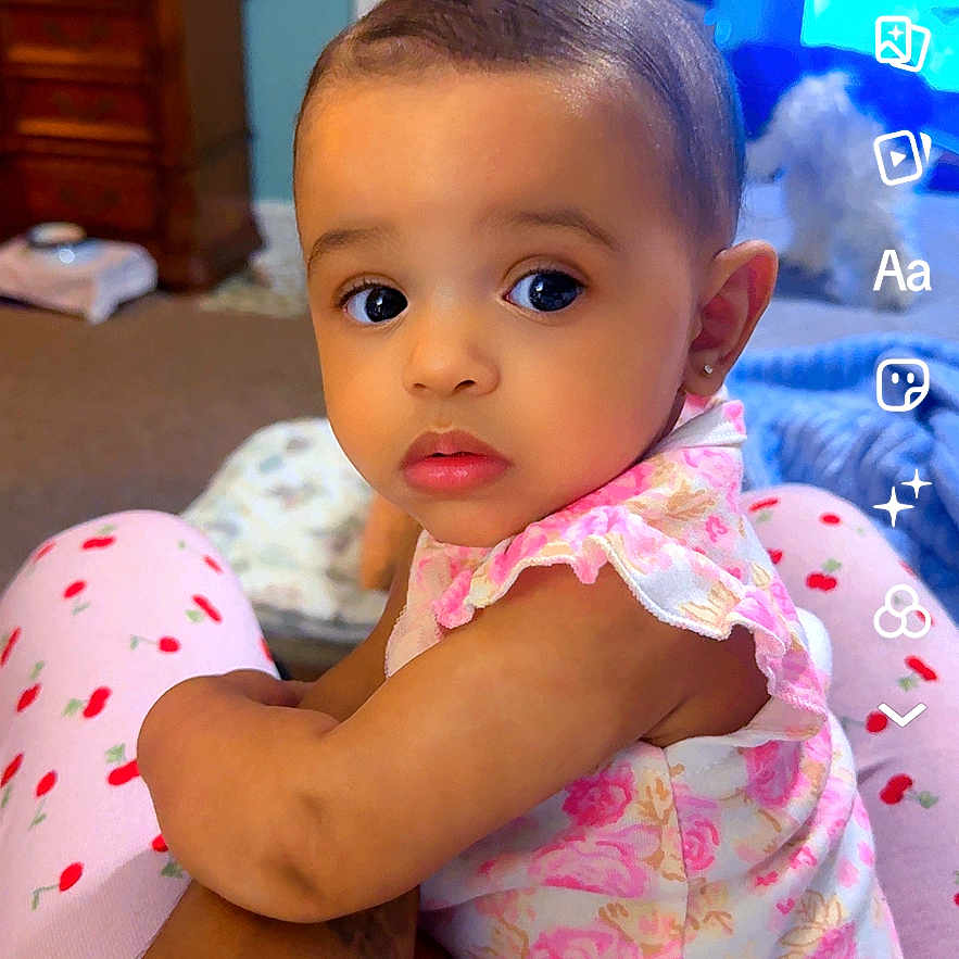 Everly is registered to the contest to win money with this photo: baby, child, infant, pink_clothing, floral, person, indoor, home, furniture, carpet, arm, face, eyes, earrings, sitting, curious, digital_interface, screenshot, mobile_app, room