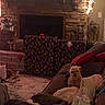 dog, couch, blanket, pillow, fireplace, stone_wall, christmas_lights, garland, wrapped_presents, rug, cozy, indoor, holiday_decor, festive, living_room, small_dog, furniture, soft_lighting, home_decor, relaxing