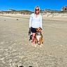 woman, dog, beach, sand, sunny, blue_sky, sunglasses, smiling, outdoor, pet, happy, casual_clothing, footwear_off, leash, dunes, residential_houses, daytime, shadow, nature, vacation