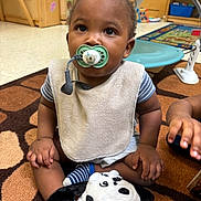 Kingston is registered to the contest to win money with this photo: toddler, pacifier, plush_toy, carpet, indoor, child, sitting, bib, striped_clothing, hand, toy, classroom, furniture, floor, play, cute, baby, expression, young_child, person