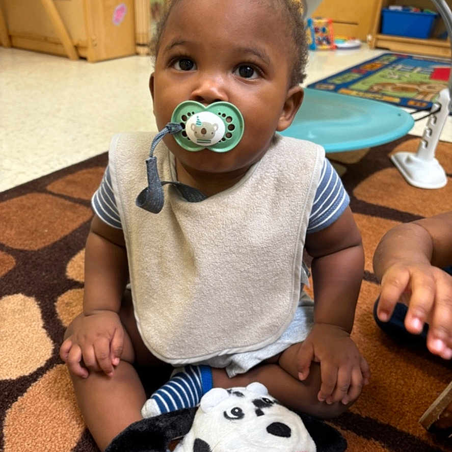 Kingston is registered to the contest to win money with this photo: baby, bib, carpet, child, classroom, cute, expression, floor, furniture, hand, indoor, pacifier, person, play, plush_toy, sitting, striped_clothing, toddler, toy, young_child