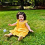 child, toddler, yellow_dress, curly_hair, grass, outdoor, sunlight, smiling, happy, nature, greenery, sitting, playful, tree, park, daylight, cute, person, footwear, flower_clip