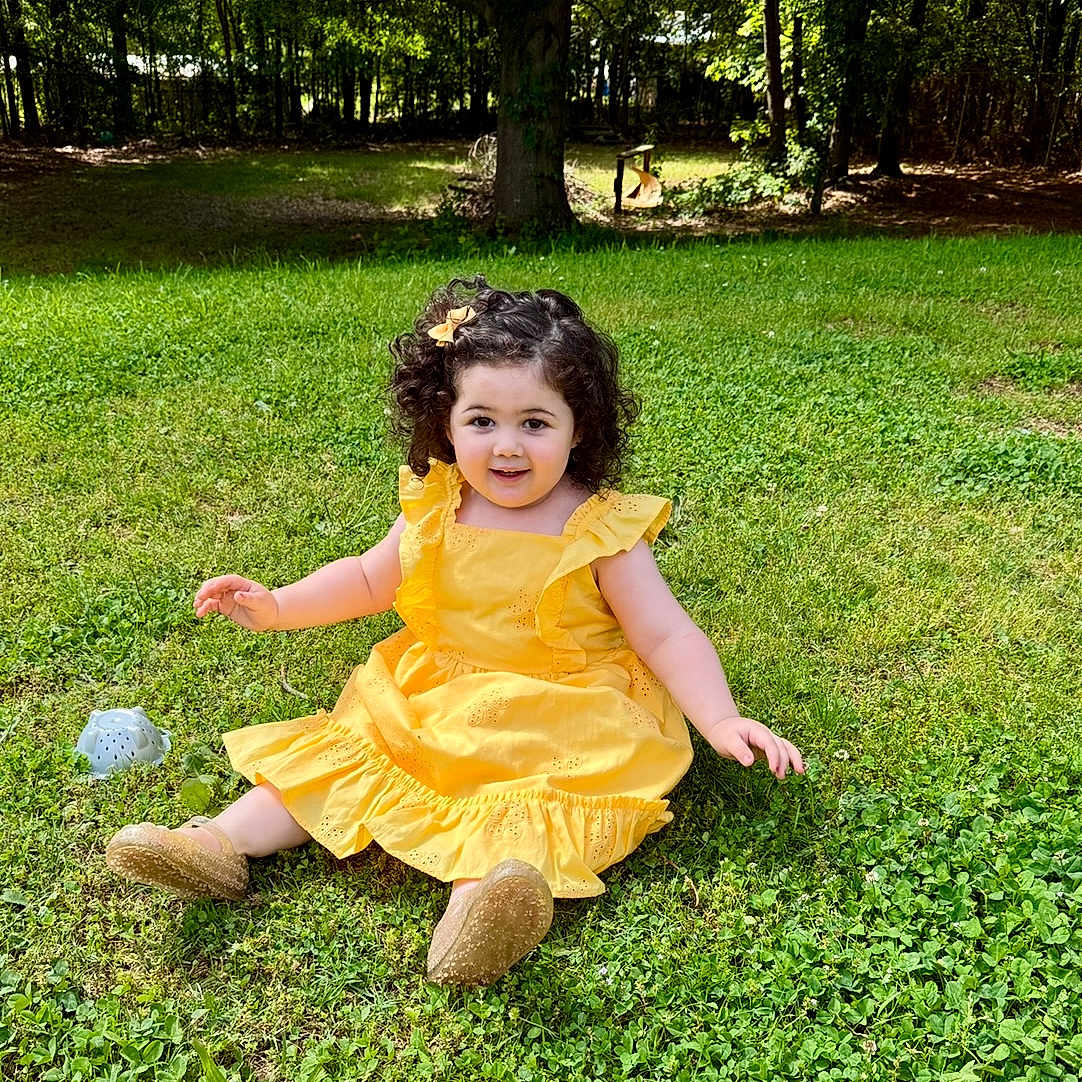 Eva joined the competition — help win amazing prizes! child, curly_hair, cute, daylight, flower_clip, footwear, grass, greenery, happy, nature, outdoor, park, person, playful, sitting, smiling, sunlight, toddler, tree, yellow_dress
