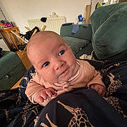 Flora is registered to the contest to win money with this photo: baby, infant, blue_eyes, blanket, giraffe_pattern, couch, living_room, socks, furniture, person, clothing, hand, head, face, portrait, indoor, wooden_floor, radiator, table, remote_control