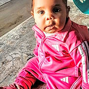 Sophia is registered to the contest to win money with this photo: baby, child, pink_tracksuit, sitting, indoor, curious, face, hair_bows, rug, shoe, footwear, laundry_basket, floor, expression, portrait, casual_clothing, young_child, person, home, cute