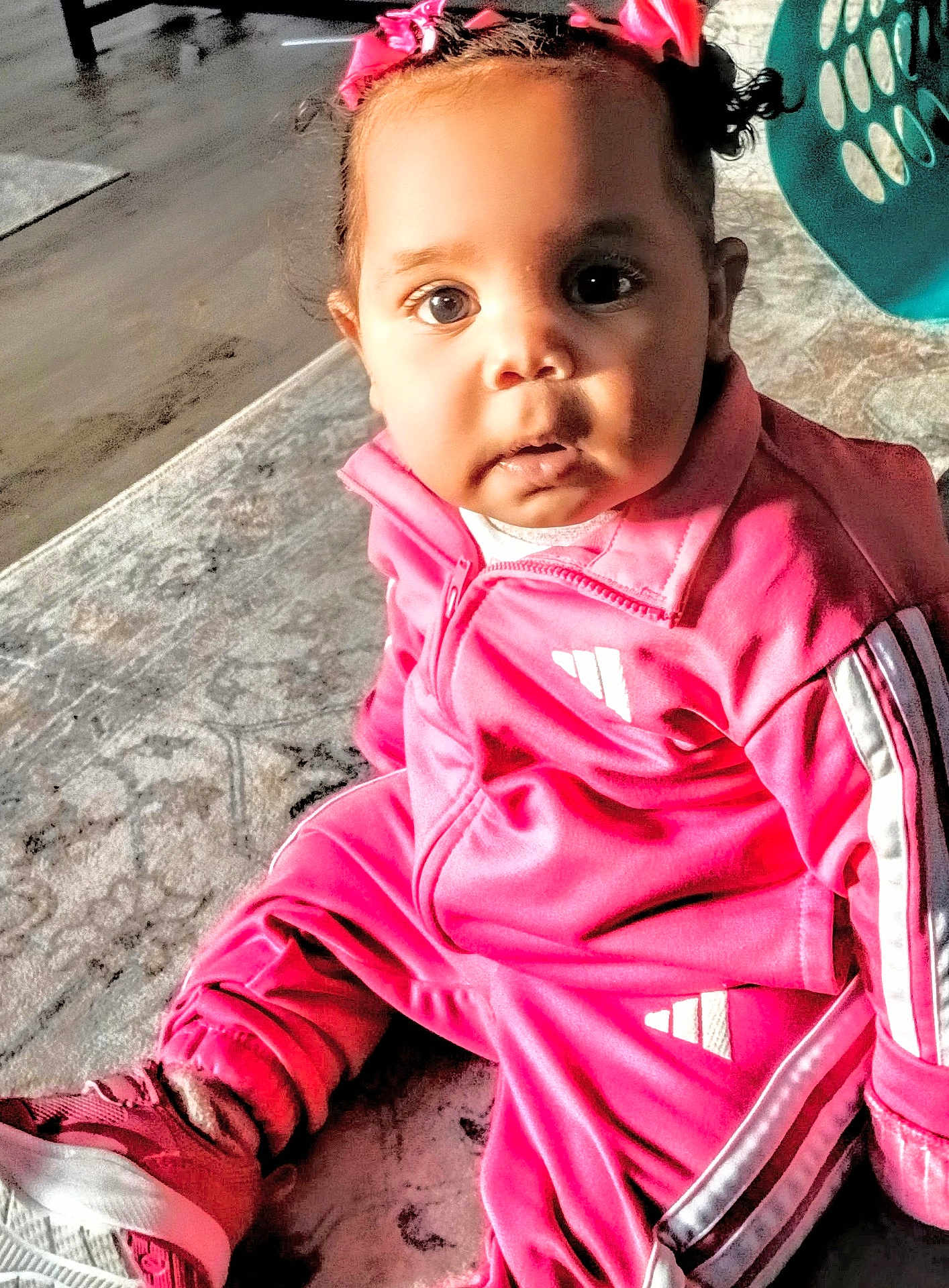 Sophia is registered to the contest to win money with this photo: baby, child, pink_tracksuit, sitting, indoor, curious, face, hair_bows, rug, shoe, footwear, laundry_basket, floor, expression, portrait, casual_clothing, young_child, person, home, cute