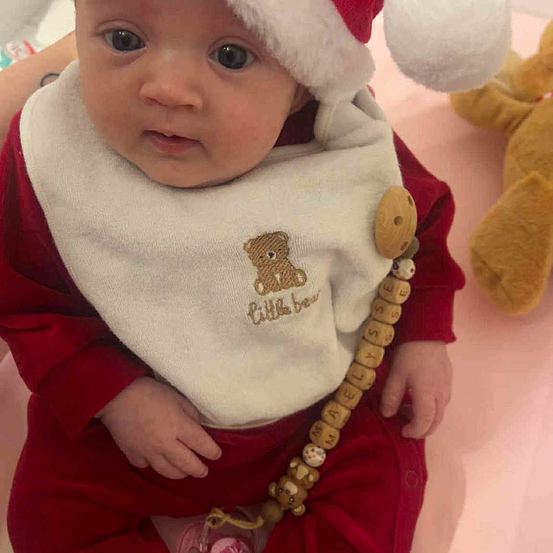 Maëlysse a rejoint le concours — aidez-le/la à gagner de superbes lots ! baby, bib, child, christmas, cozy, cute, face, headwear, holiday, indoors, infant, pacifier, pacifier_clip, person, plush_toy, red_clothing, santa_hat, sitting, soft_toy, teddy_bear