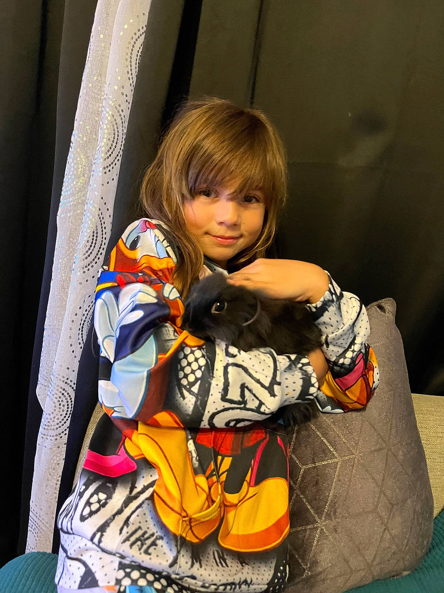animal, black_bunny, bunny, child, colorful_clothing, couch, cuddling, curtain, cute, furry, home, hoodie, indoor, person, pet, pillow, portrait, relaxed, smile, young