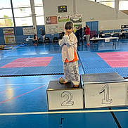 Louane a rejoint le concours — aidez-le/la à gagner de superbes lots ! child, taekwondo, martial_arts, podium, gym, sports_uniform, barefoot, blue_floor, competition, people, indoor, sport_event, young_child, orange_shirt, white_uniform, medal_stand, pose, person, mat, victory