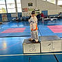 child, taekwondo, martial_arts, podium, gym, sports_uniform, barefoot, blue_floor, competition, people, indoor, sport_event, young_child, orange_shirt, white_uniform, medal_stand, pose, person, mat, victory