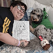Mylo joined the competition — help win amazing prizes! bed, black_shirt, blanket, coloring_book, curly_hair, dog, drawing, glasses, green_clothing, hoodie, indoor, lip_piercing, nose_ring, patterned, person, pet, piercing, plush_toy, smile, tattoo