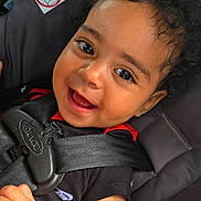 Maddox is registered to the contest to win money with this photo: baby, child, smile, happy, car_seat, seat_belt, curly_hair, black_shirt, infant, closeup, face, cute, young_child, safety, car_safety, patterned_blanket, indoors, skin, person, portrait