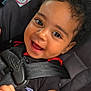 baby, child, smile, happy, car_seat, seat_belt, curly_hair, black_shirt, infant, closeup, face, cute, young_child, safety, car_safety, patterned_blanket, indoors, skin, person, portrait