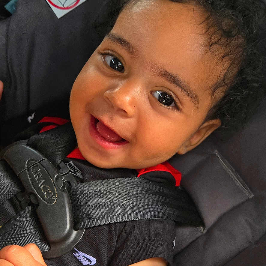 Maddox is registered to the contest to win money with this photo: baby, black_shirt, car_safety, car_seat, child, closeup, curly_hair, cute, face, happy, indoors, infant, patterned_blanket, person, portrait, safety, seat_belt, skin, smile, young_child