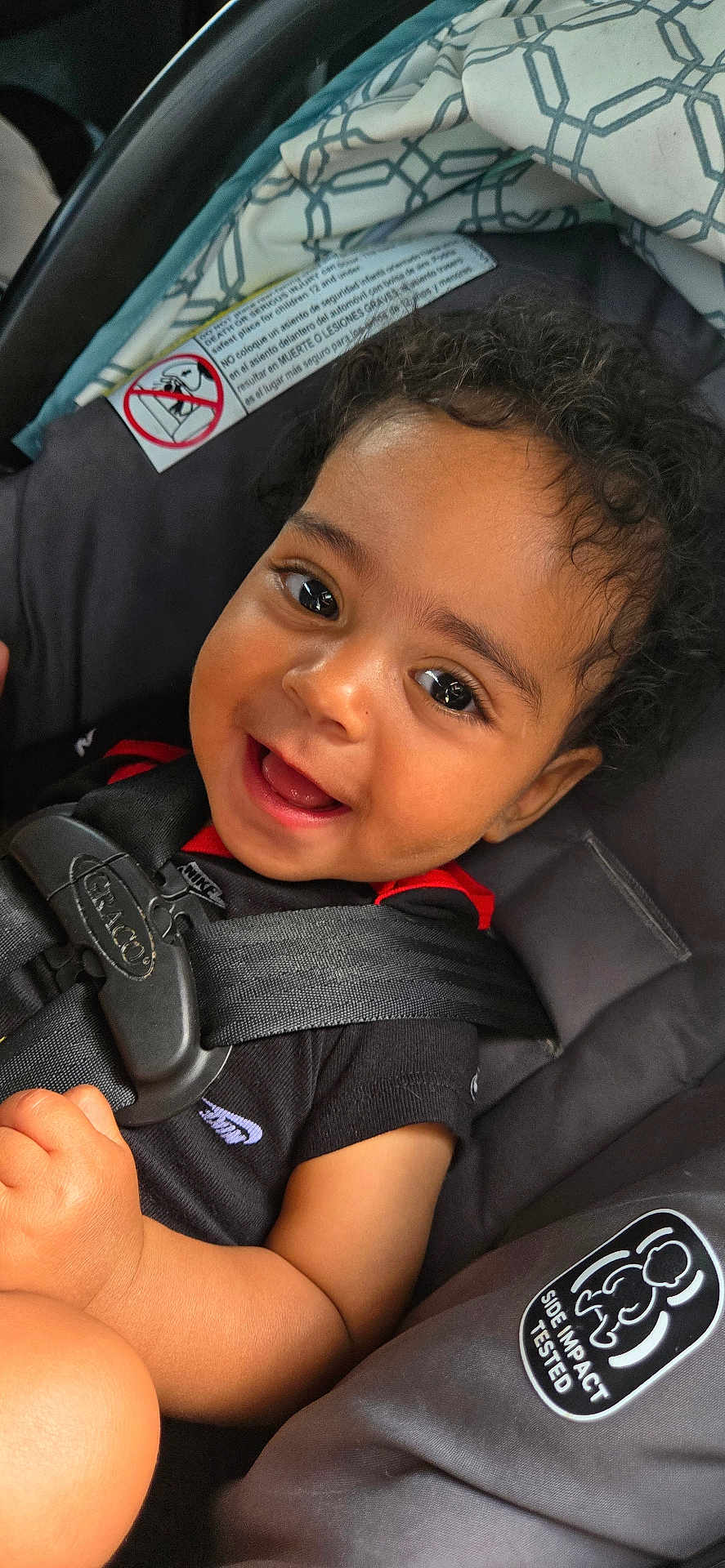 Maddox is registered to the contest to win money with this photo: baby, child, smile, happy, car_seat, seat_belt, curly_hair, black_shirt, infant, closeup, face, cute, young_child, safety, car_safety, patterned_blanket, indoors, skin, person, portrait