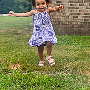 Camila is registered to the contest to win money with this photo: child, toddler, girl, smile, happy, running, dress, purple, floral_pattern, sandals, grass, outdoor, curly_hair, brick_wall, greenery, arms_outstretched, playful, daylight, person, nature