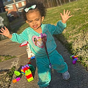 Camila is registered to the contest to win money with this photo: arms_raised, bow, building_blocks, child, curly_hair, facial_expression, grass, outdoor, playground, playtime, portrait, residential_background, sidewalk, smiling, sneakers, standing, sunlight, toddler, toys, turquoise_clothing