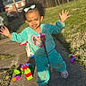 arms_raised, bow, building_blocks, child, curly_hair, facial_expression, grass, outdoor, playground, playtime, portrait, residential_background, sidewalk, smiling, sneakers, standing, sunlight, toddler, toys, turquoise_clothing