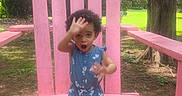 Camila joined the competition — help win amazing prizes! child, toddler, blue_clothing, heart_pattern, pink_chair, outdoor, wood, tree, grass, playful, kneeling, curly_hair, daylight, nature, expression, fun, park, summer, wooden_furniture, person
