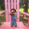 child, toddler, blue_clothing, heart_pattern, pink_chair, outdoor, wood, tree, grass, playful, kneeling, curly_hair, daylight, nature, expression, fun, park, summer, wooden_furniture, person