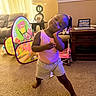 toddler, child, indoor, living_room, carpet, play_tent, barney, furniture, lamp, table, sofa, curtains, hair_braids, pink_shirt, shorts, barefoot, smile, pose, toy, home