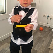 Noam a rejoint le concours — aidez-le/la à gagner de superbes lots ! child, toddler, boy, tuxedo, bow_tie, vest, white_shirt, black_pants, barefoot, indoors, wooden_floor, window, orange_ball, electrical_outlets, curious_expression, holding_object, standing, person, clothing, floor
