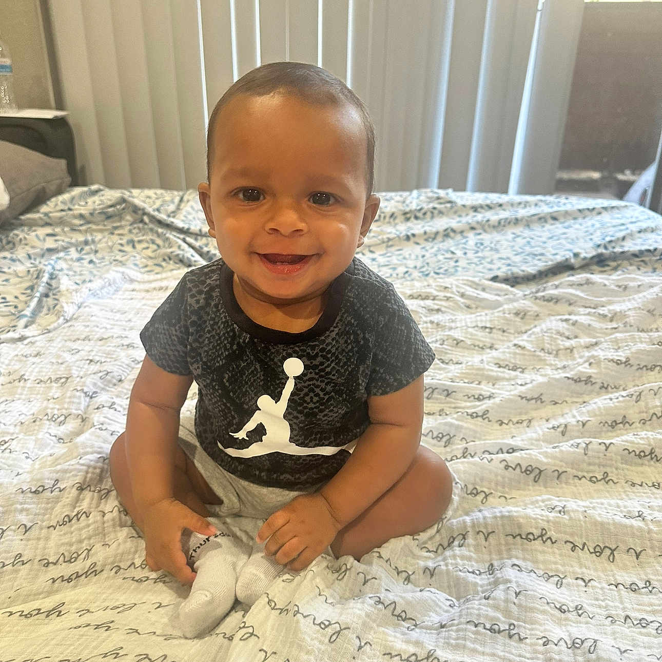 Jaxon is registered to the contest to win money with this photo: baby, child, smiling, sitting, bed, blanket, indoor, vertical_blinds, clothing, shorts, shirt, person, face, happy, infant, skin, footwear, sock, home, daylight