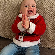 Roman is registered to the contest to win money with this photo: baby, child, clapping, red_sweater, santa_theme, jeans, couch, beige_cushion, happy, smiling, indoor, cute, holiday, festive, portrait, seated, infant, cozy, warm_clothing, expression