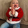 baby, child, clapping, red_sweater, santa_theme, jeans, couch, beige_cushion, happy, smiling, indoor, cute, holiday, festive, portrait, seated, infant, cozy, warm_clothing, expression