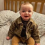 Roman is registered to the contest to win money with this photo: baby, smiling, corduroy_jacket, pants, bed, pillow, wooden_headboard, happy, child, infant, clothing, person, indoor, portrait, sitting, cute, face, hands, footwear, textile