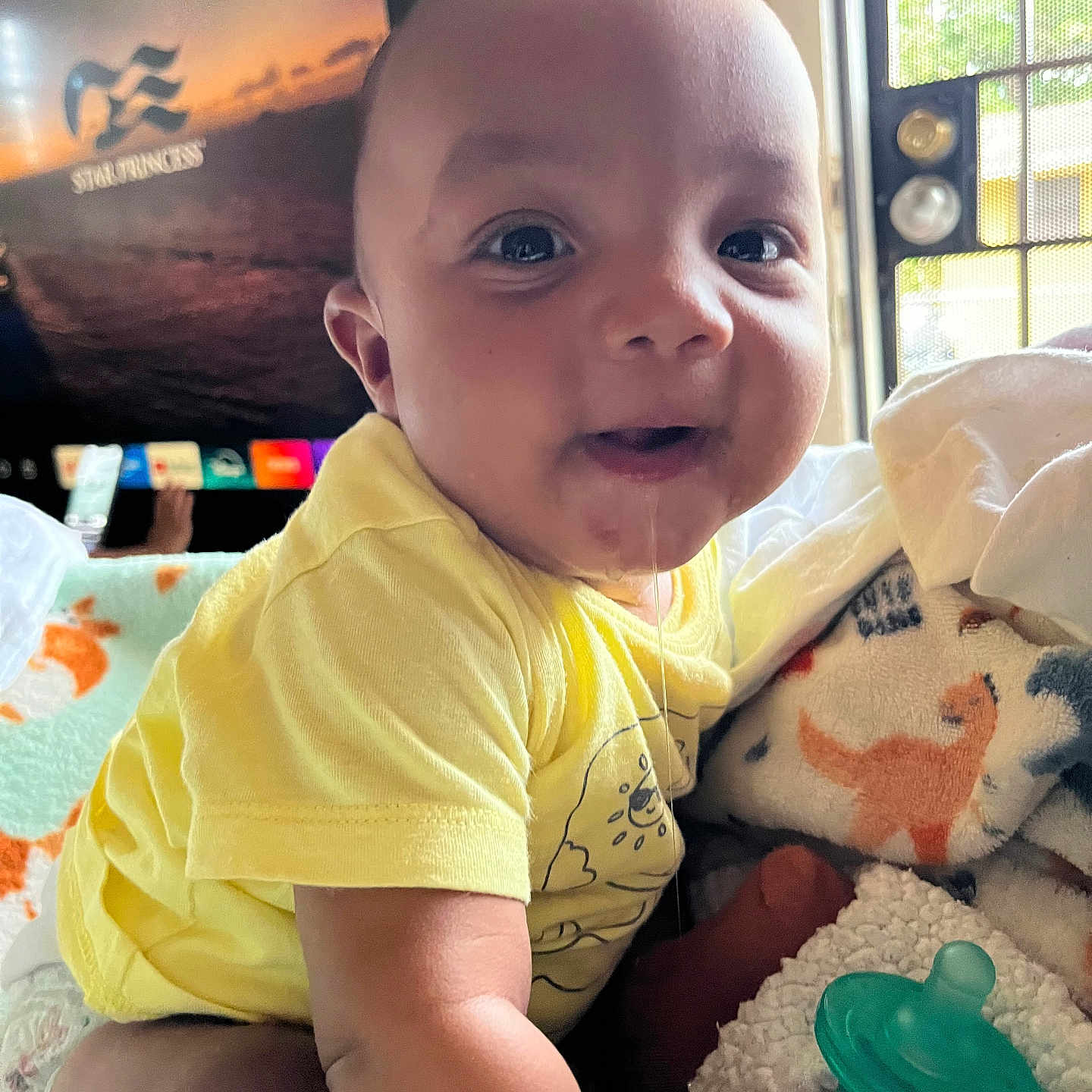 Tynan is registered to the contest to win money with this photo: baby, child, infant, smiling, happy, portrait, yellow_outfit, sitting, blanket, plush_toy, pacifier, toy, bedroom, indoors, window, television, home, sunlight, close_up, face