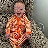 Michael is registered to the contest to win money with this photo: baby, child, infant, smiling, orange_striped_clothing, onesie, happy, sitting, couch, patterned_furniture, indoor, face, feet, footwear, pumpkin_design, cute, joyful, person, home, cozy