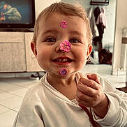 Louna a rejoint le concours — aidez-le/la à gagner de superbes lots ! child, toddler, smiling, stickers_on_face, sticker, indoors, living_room, television, cabinet, tile_floor, hand, sweater, portrait, cute, eyes, teeth, hair, ears, playful, pink_sticker
