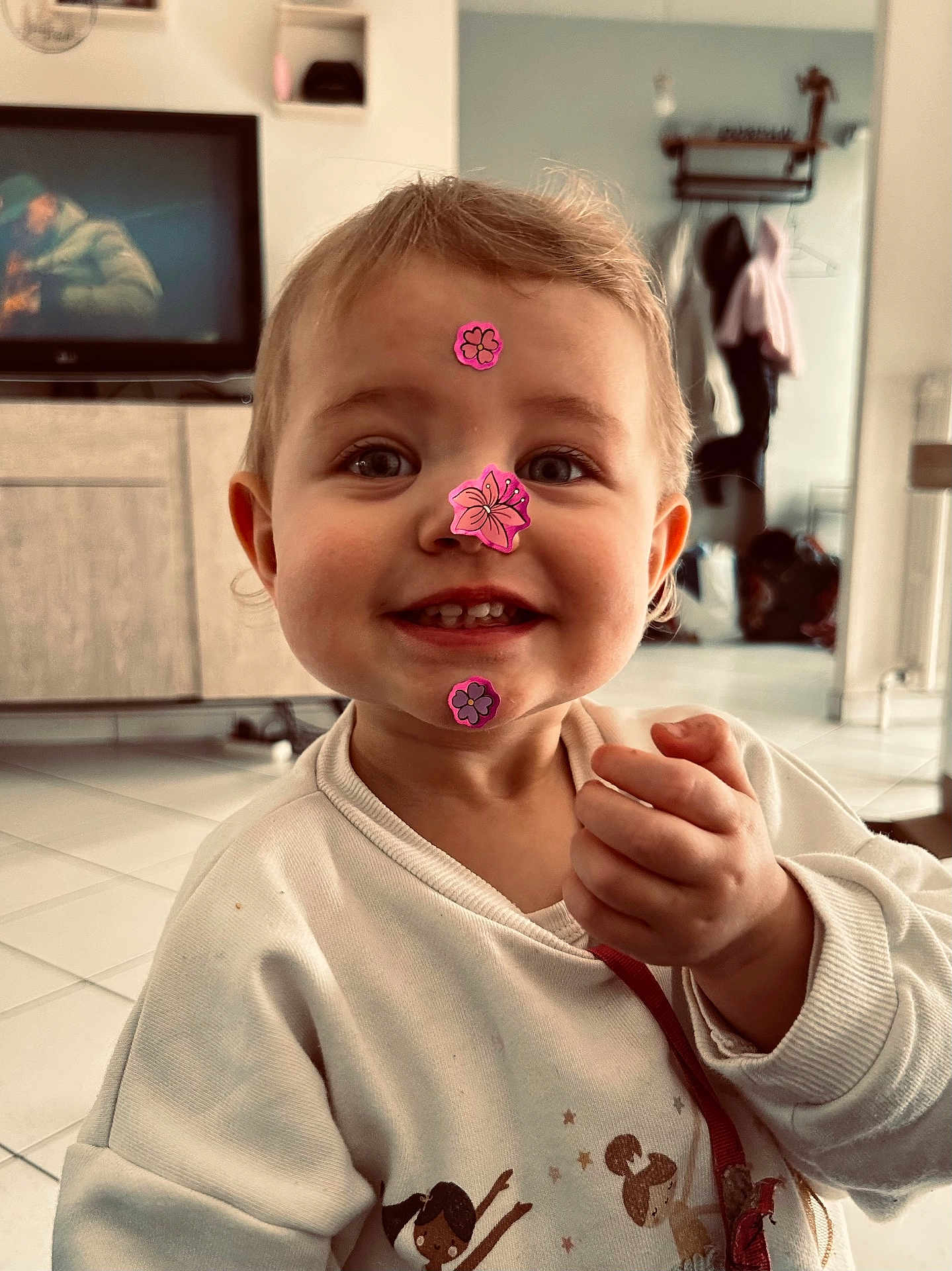 Louna a rejoint le concours — aidez-le/la à gagner de superbes lots ! child, toddler, smiling, stickers_on_face, sticker, indoors, living_room, television, cabinet, tile_floor, hand, sweater, portrait, cute, eyes, teeth, hair, ears, playful, pink_sticker