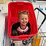 child, smiling, laundromat, washing_machine, laundry_cart, red_cart, toddler, plaid_pants, black_and_white_shirt, floor, metal_cart, yellow_container, indoor, happy, person, toy_cart, hair, face, clothing, laundry_basket