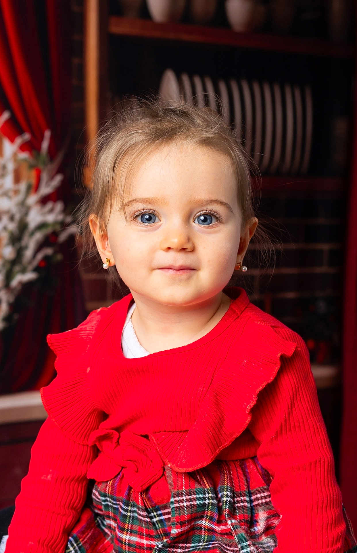 Margot a rejoint le concours — aidez-le/la à gagner de superbes lots ! child, toddler, girl, portrait, blue_eyes, red_clothing, plaid_skirt, earrings, smile, indoor, festive, holiday, bookshelf, plates, curtain, close_up, face, hair, eyelashes, sitting