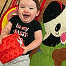 baby, black_shirt, carpet, child, colorful, cute, happy, indoor, infant, joyful, lying_down, panda, person, playful, red_block, smiling, soft_toy, text_on_clothing, toy, young_child