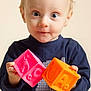 toddler, child, blonde_hair, blue_eyes, toy_blocks, pink_block, orange_block, navy_shirt, indoor, portrait, curious, holding, baby, cute, face, hands, playing, young_child, studio_lighting, soft_background