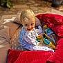 toddler, child, pajamas, smiling, toys, red_pillows, beige_pillows, cushions, indoor, playtime, happy, blond_hair, blue_eyes, person, sitting, portrait, cozy, soft_lighting, home, cute
