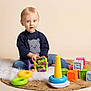 toddler, child, toy, colorful, sitting, blue_jeans, sweater, indoor, rug, plastic_toys, blocks, stacking_rings, fur_rug, expression, portrait, playtime, cute, studio, light_background, floor