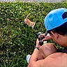 Sunny is registered to the contest to win money with this photo: person, cat, kitten, grass, outdoor, camera, photography, blue_cap, greenery, nature, animal, pet, young_cat, crouching, summer, daylight, cute, curious, exploring, wildflowers