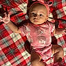 baby, infant, child, portrait, lying_down, pink_outfit, headband, bow, socks, plaid_blanket, red_blanket, indoor, close_up, face, eyes, arms_raised, smiling, fashion, textured_fabric, cute